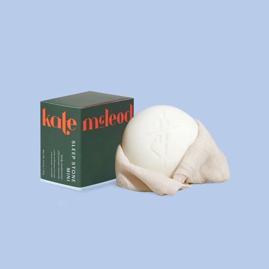 A round KATE MCLEOD - SLEEP STONE MINI LOTION BAR 1oz sits on lavender chamomile sprigs with a light blue backdrop. Above, the text reads "Soothing Herbal.
