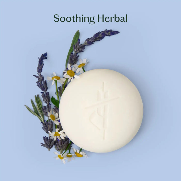 A round, white KATE MCLEOD - SLEEP STONE MINI LOTION BAR 1OZ rests partly wrapped in beige cloth beside a green box with orange “kate mcleod Sleep Stone Mini” lettering, infused with lavender chamomile, on a light blue background.