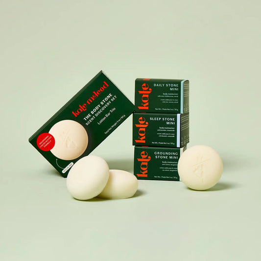 The KATE MCLEOD SCENT DISCOVERY SET features three mini, round white lotion bars with a plant-based formula. The bars come in green and orange branded packaging, stacked beside a larger box on a light green background.