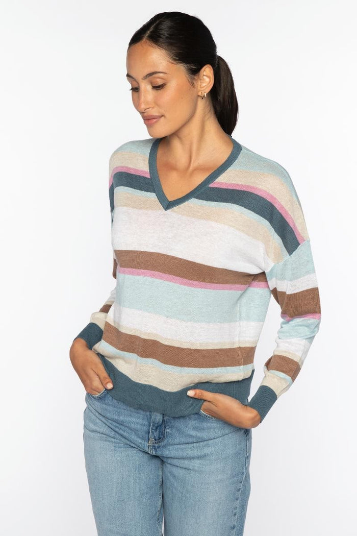 A woman with dark hair in a ponytail wears the KINROSS CASHMERE Multi Color Stripe Vee Neck Linen Sweater and blue jeans, standing with her hands in her pockets and smiling slightly against a white background.