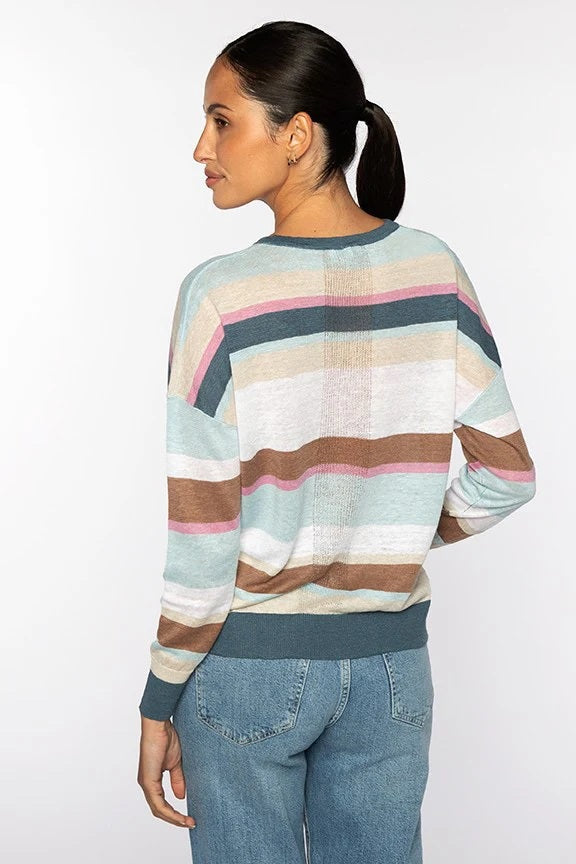 A woman with dark hair in a ponytail wears the KINROSS CASHMERE Multi Color Stripe Vee Neck Linen Sweater and blue jeans, standing with her hands in her pockets and smiling slightly against a white background.