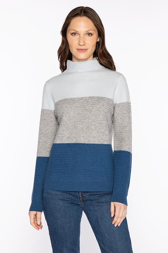 A woman wearing a color-blocked turtleneck sweater in light blue, gray, and dark blue, paired with blue jeans, stands against a plain white background.