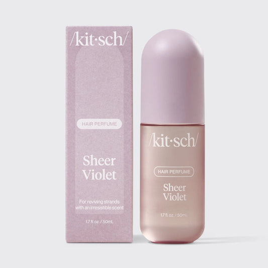 A 1.7 fl oz bottle of KITSCH - HAIR PERFUME MIST IN SHEER VIOLET sits beside its coordinating lavender box against a light gray background. Both the bottle and box feature the product and brand name in white text, emphasizing this sophisticated hair perfume mist from KITSCH, characterized by floral-fruity-musk notes and advanced odor-eliminating technology.