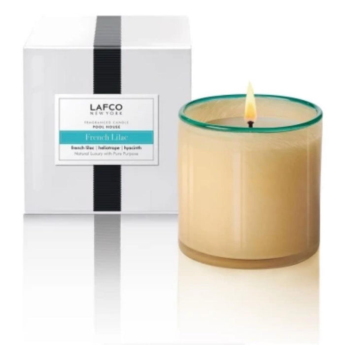 A lit LAFCO French Lilac Poolhouse 15oz luxury candle in a glass holder sits before its white box, featuring a black vertical stripe and a label that matches the product name.