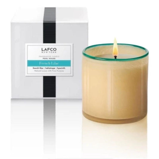 A lit LAFCO French Lilac Poolhouse 15oz luxury candle in a glass holder sits before its white box, featuring a black vertical stripe and a label that matches the product name.