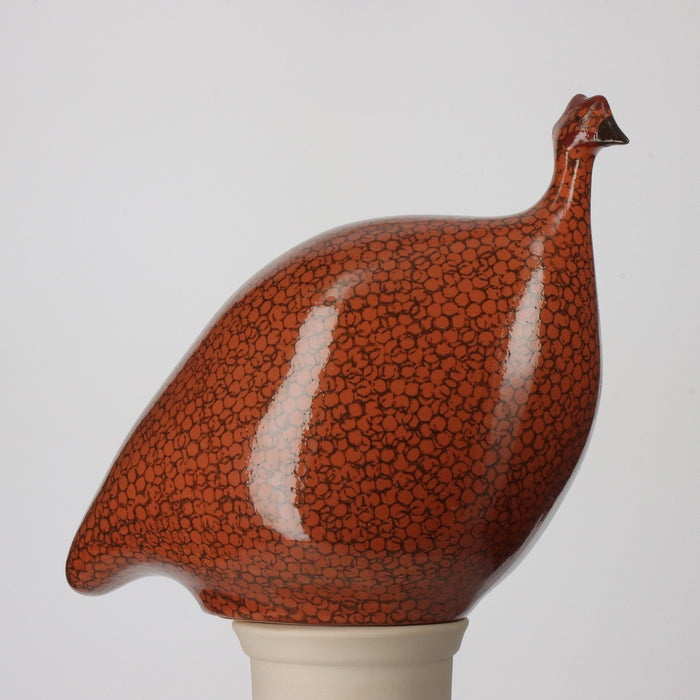 The LES CERAMIQUES DE LUSSAN - Medium Ceramic French Guinea in terracotta red with black spots features a rounded body and small head, handmade in the South of France and displayed on a white cylindrical stand.