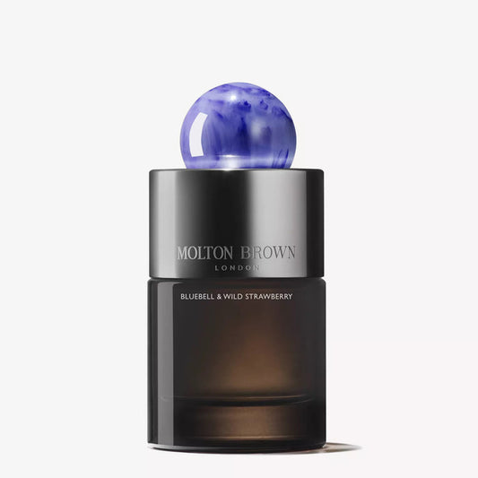 Three Molton Brown Bluebell & Wild Strawberry Eau de Parfum bottles sit on a blue surface with fresh strawberries, emphasizing the collection’s wild strawberry and floral-fruity scent. A silver tube is placed on the right side.