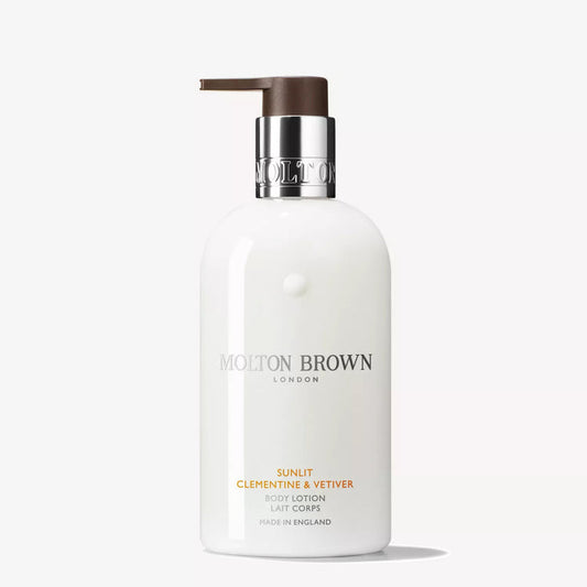 Three Molton Brown bath and body products—including the Sunlit Clementine & Vetiver Body Lotion—plus a rectangular box sit on a neutral surface with a small card, set against a warm mustard-yellow background.