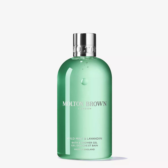 A set of MOLTON BROWN toiletries, featuring the WILD MINT AND LAVANDIN BATH & SHOWER GEL, a rollerball vial, a jar with a green sphere, and a small tin, displayed on mossy bark with a green and beige background.