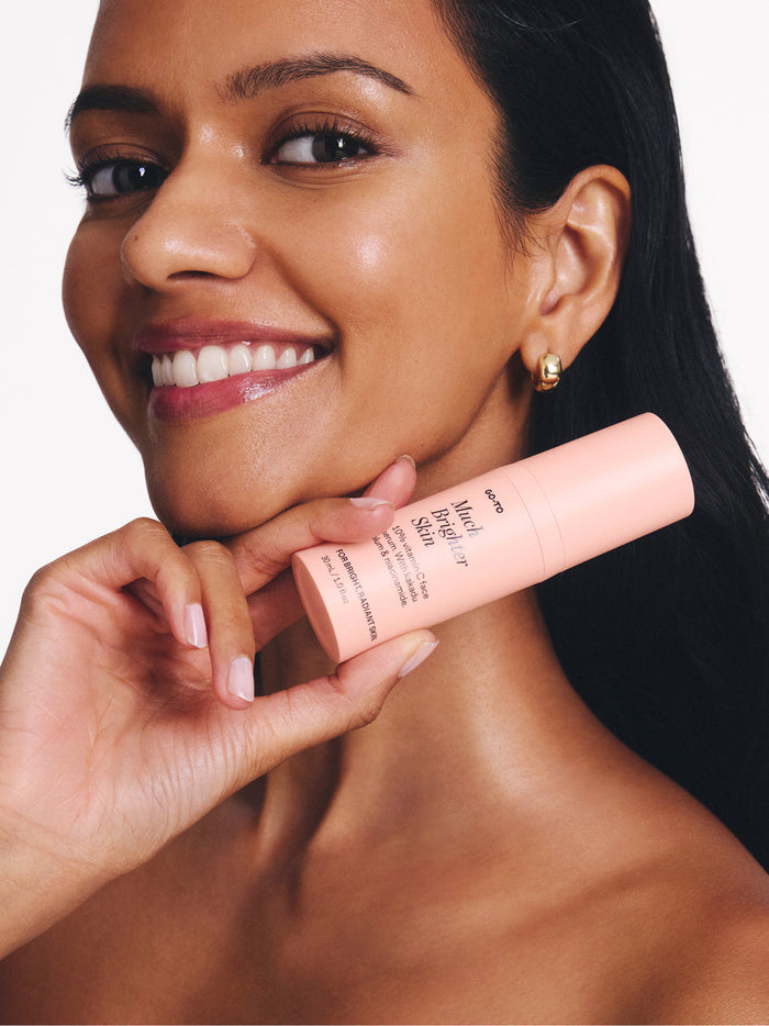 Smiling woman holds a pink skincare bottle labeled "brighter skin," showcasing glowing complexion and gold earrings