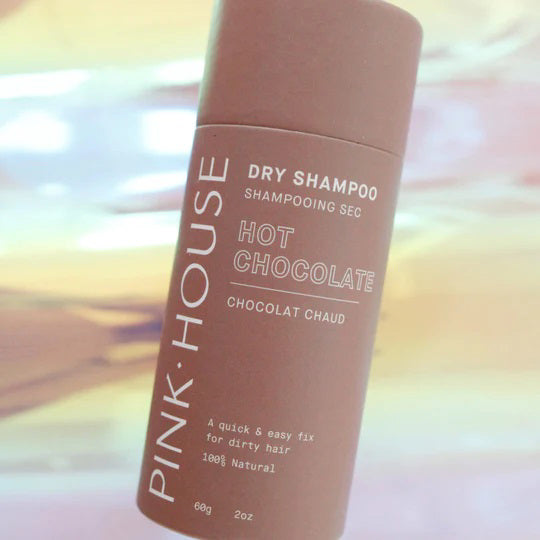 A cylindrical PINK HOUSE ORGANICS - DRY SHAMPOO HOT CHOCOLATE stands upright, featuring white text on a brown background with an abstract pastel backdrop—ideal for adding volume and refreshing greasy hair.