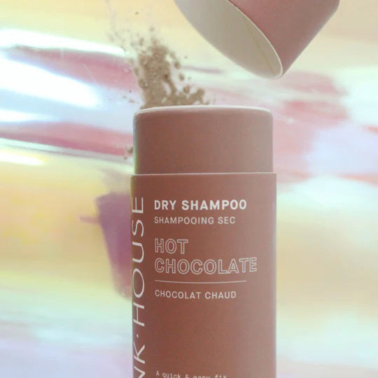 A cylindrical PINK HOUSE ORGANICS - DRY SHAMPOO HOT CHOCOLATE stands upright, featuring white text on a brown background with an abstract pastel backdrop—ideal for adding volume and refreshing greasy hair.