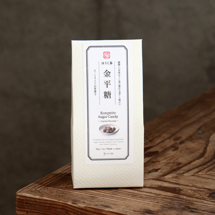 A white rectangular package of Japanese konpeito sugar candy stands upright on a rustic wooden surface against a dark gray background.